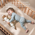 A smiling baby in light blue ribbed pajamas lies on a cream-colored crib mattress, hugging the soft otter sensory toy that doubles as a measuring tool. The toy is stretched out alongside the baby, showing its full length with measurement indicators clearly visible on the underside. The plush toy features a brown otter head at one end and colorful segments between, with measurement markings ranging up to 40cm as indicated by text at the bottom of the image. The blue whale teether is visible with a zigzag lin