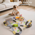 A young child playing on a carpet with an adventure dinosaur track toy set that includes looping tracks, a dinosaur head tunnel, small cars, plastic palm trees, and dinosaur figurines. The scene is in a cozy living room with text at the top that says 