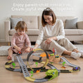 A mother and child sitting on the floor in a living room, happily playing together with a dinosaur-themed race track toy set that features looping tracks, a green dinosaur head tunnel, toy cars, palm trees, and dinosaur figures.